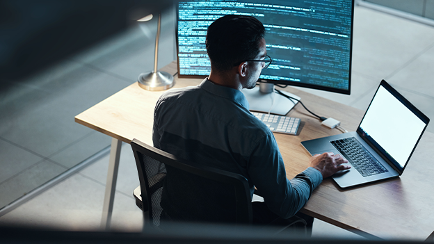 Photograph of a man sat at a desk using a laptop computer and a larger desktop monitor